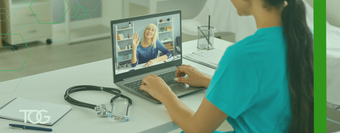 A female doctor with long brown hair dressed in green scrubs has a telemedicine visit with a patient using her laptop.