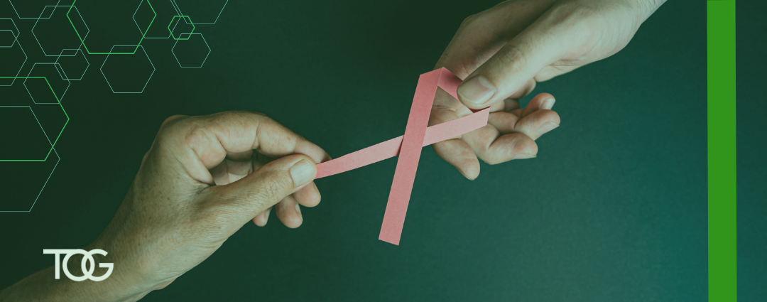 Two hands coming together against a dark green background to hold a pink ribbon symbolizing breast cancer awareness.
