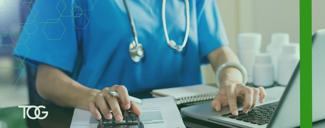 Faceless doctor wearing blue scrubs and a stethoscope around their neck enters data into a calculator in front of a laptop computer.