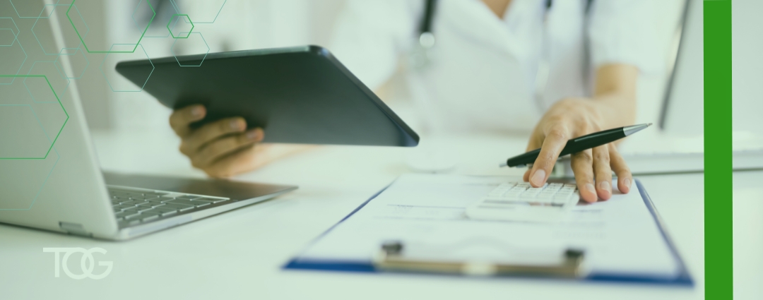 A healthcare professional uses a calculator while reviewing documents on a clipboard, with a tablet and laptop on the desk.