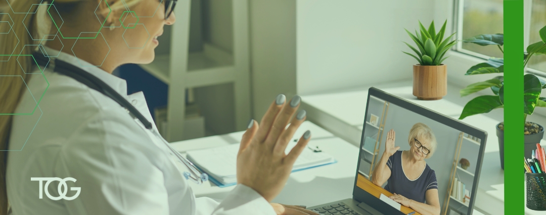 A healthcare professional wearing a stethoscope waves during a video consultation with an older woman on a laptop screen, in a bright office with plants by the window.