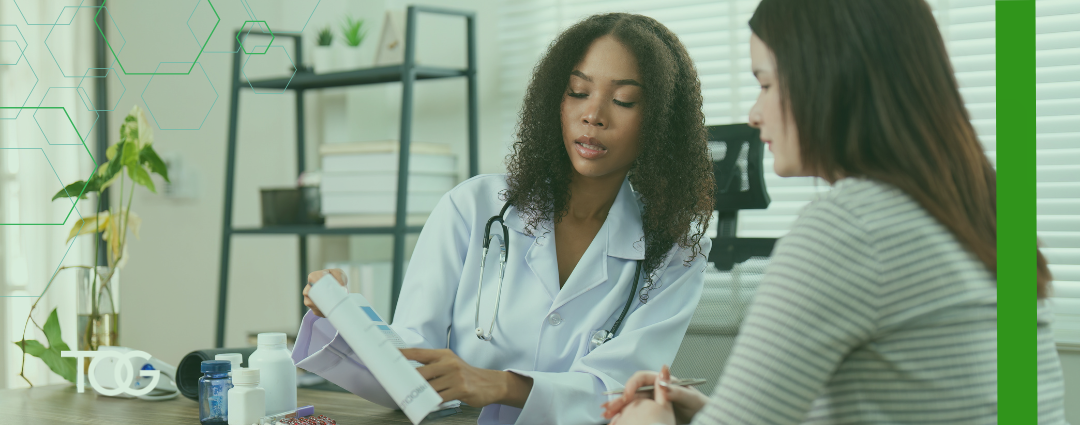 Female doctor with shoulder length curly brown hair wearing a white lab coat sits at table with female patient to review prescription instructions.
